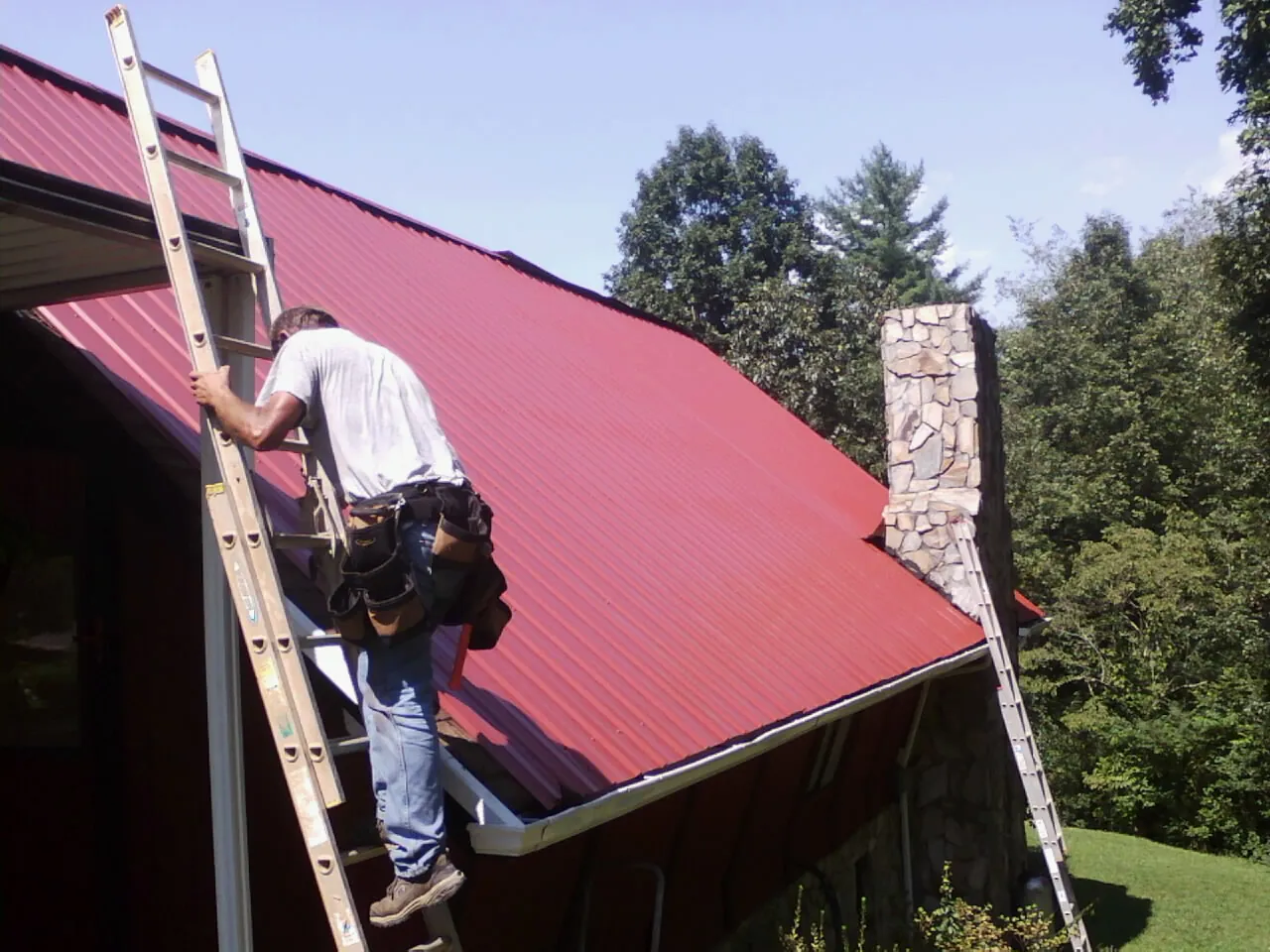 Roofer on a ladder beside a red metal roof and stone chimney — Soffit & Fascia Repair by Roofing Brothers in Philomath